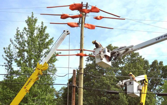A worker in a bucket truck repairs an overhead power line, with another worker visible on the ground. The image illustrates the need for robust broadband infrastructure rather than temporary solutions like hotspots.