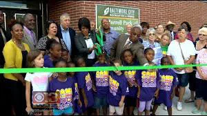 A group of people, including children in matching t-shirts and several city officials, cut a green ribbon to celebrate the opening of a community center. The event marks a potential investment opportunity for Baltimore's American Rescue Plan Act (ARPA) funds.