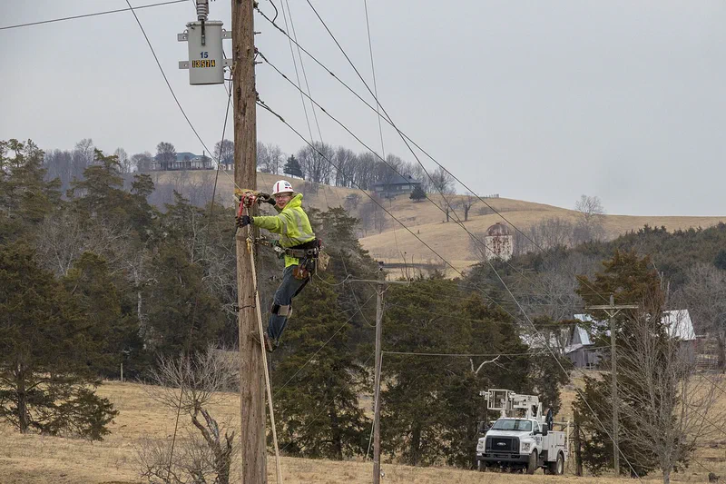 A lineman in a white hard hat and high-visibility jacket climbs a utility pole above rolling rural farmland, with farmhouses on the hill behind him and a bucket truck parked below.