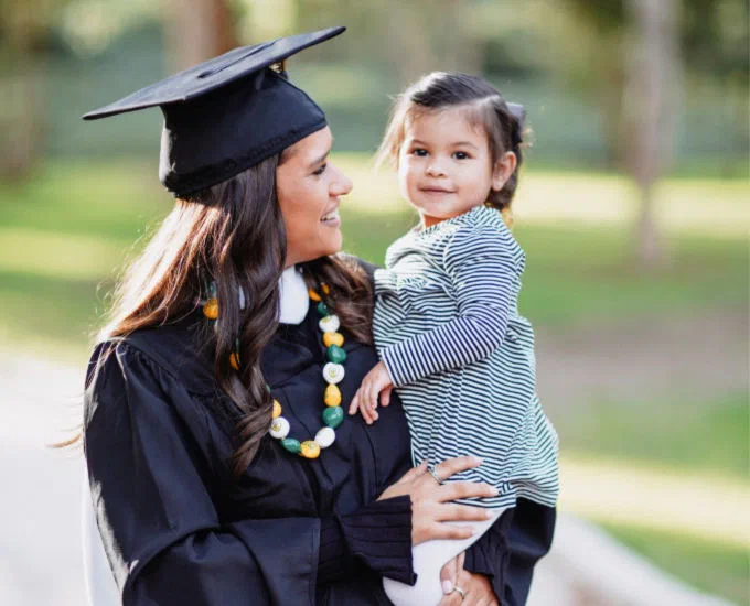 A woman in a graduation gown and cap smiles at her young daughter, who is being held in her arms. This image illustrates the challenges faced by students with families and highlights the potential benefits of financial support for community college students.