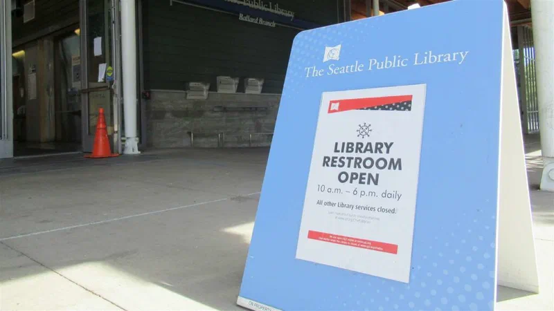 A sandwich-board sign outside the Ballard Branch of The Seattle Public Library advertises restroom availability, noting hours and that all other library services are closed. This highlights the need for maintaining public facilities like restrooms, as discussed in the article.