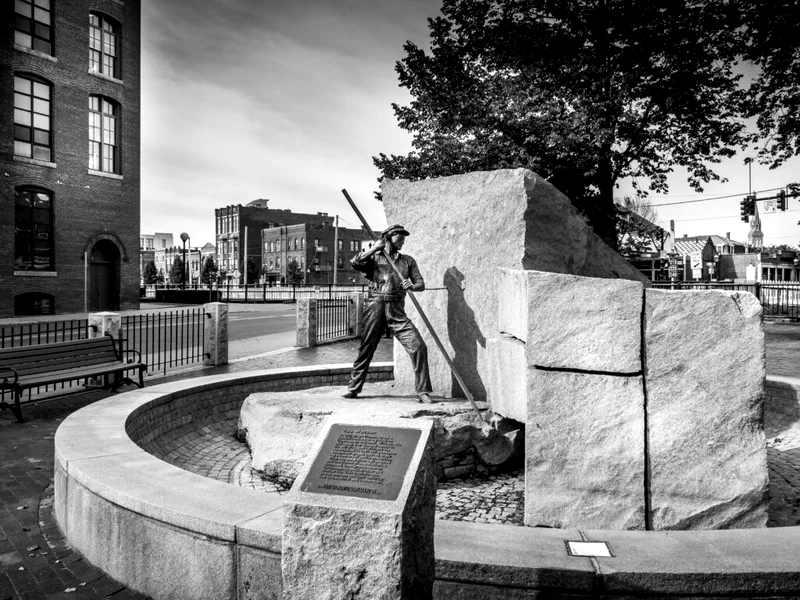 This black and white photograph depicts 'The Worker' memorial in Lowell, Massachusetts. The sculpture shows a man in working clothes leaning on a large stone block, symbolizing the city's history as a textile manufacturing hub.