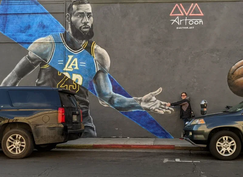 A mural of LeBron James in a Los Angeles Lakers jersey is painted on the side of a building, with a person and SUV visible in front. The image illustrates James's commitment to community investment through his I PROMISE Village and other initiatives. Jared is high-fiving the mural of LeBron.