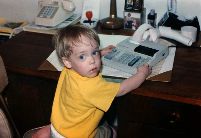 A photograph shows a toddler seated at a desk, looking back at the camera, as he play s with an old desk calculator. Jared Knowles is the toddler shown.