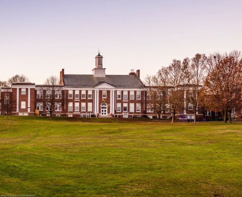 This photograph shows a large, brick school building with white columns and a central cupola, set against a backdrop of bare trees and a grassy lawn. The image illustrates the potential for accountability systems, similar to those used in schools, to be applied to police departments.