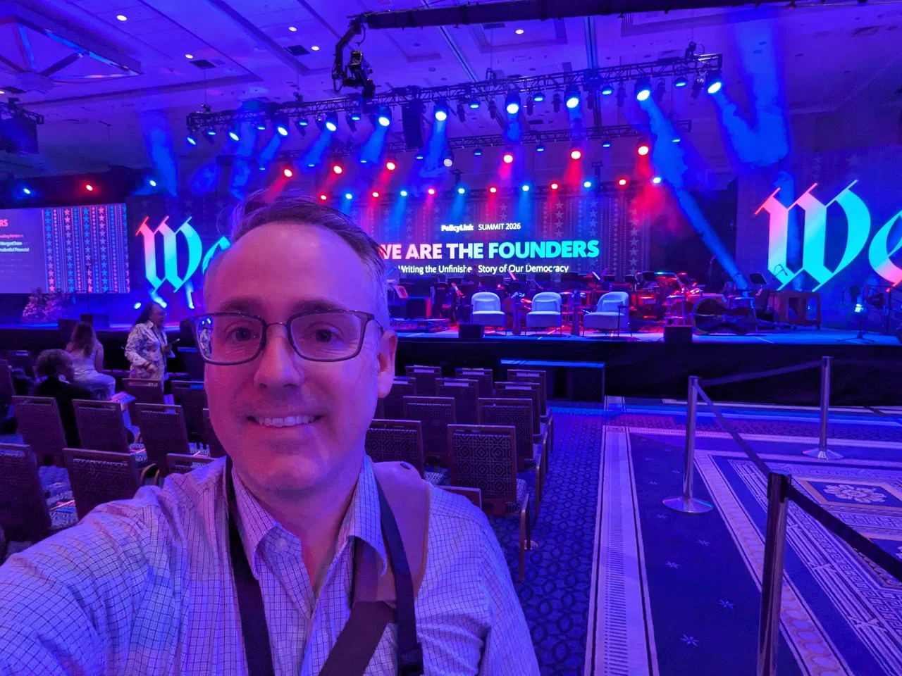 Jared Knowles takes a selfie in the main hall at PolicyLink Summit 2026. Behind him the stage is lit in blue and red, with the summit title displayed on screen: We Are the Founders, Writing the Unfinished Story of Our Democracy.