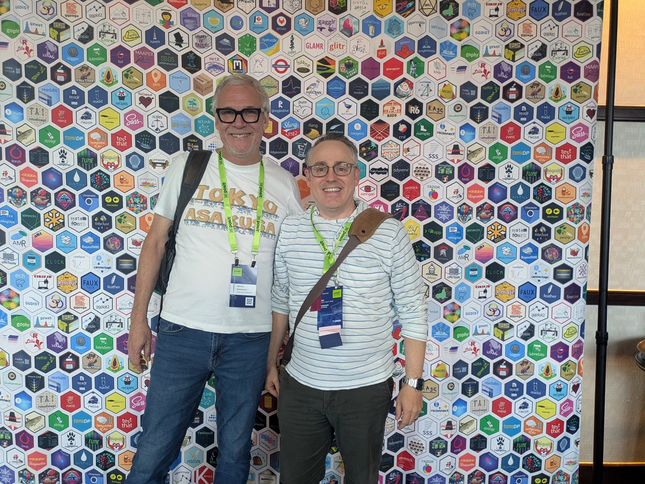 Two men stand in front of a photo backdrop of hexagons representing R package logos, the taller man on the left is Damian with Jared on the right.
