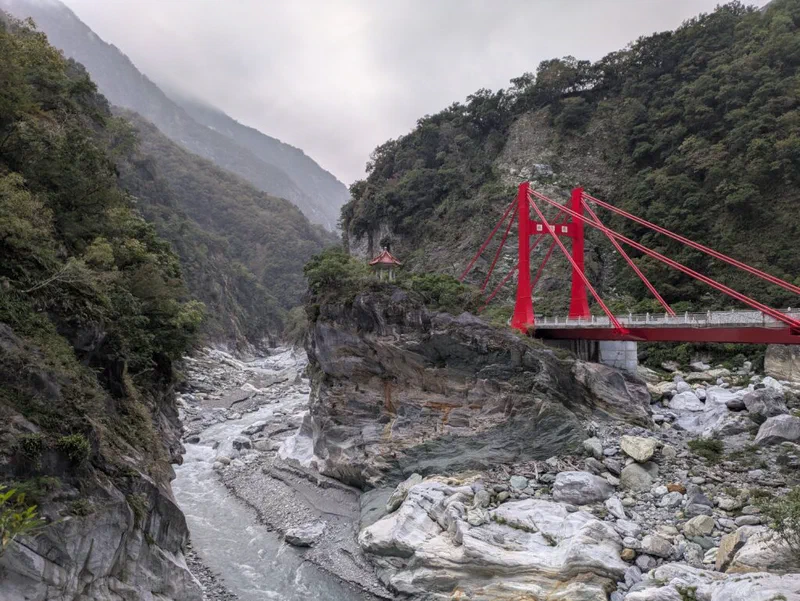 A scenic view shows a river valley in Taiwan, with a red suspension bridge spanning the water and a traditional temple visible on the hillside. The landscape highlights the natural beauty of Taiwan.