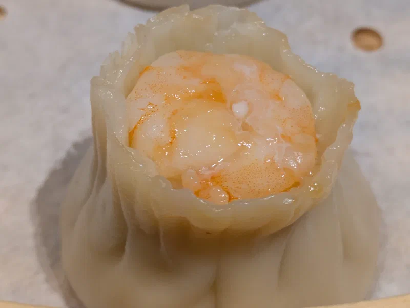 A close-up shows a single, steamed shrimp dumpling resting on a bamboo steamer. The dumpling's translucent wrapper encases a bright orange-red shrimp, illustrating the delicate balance local governments face in managing limited resources.