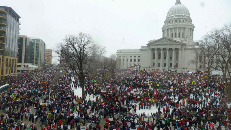 A wide photograph shows a large crowd of protestors gathered in front of the Wisconsin State Capitol building on a snowy day. The demonstration, which occurred in 2011, reflects disagreements over state preemption, labor costs, and devolution.