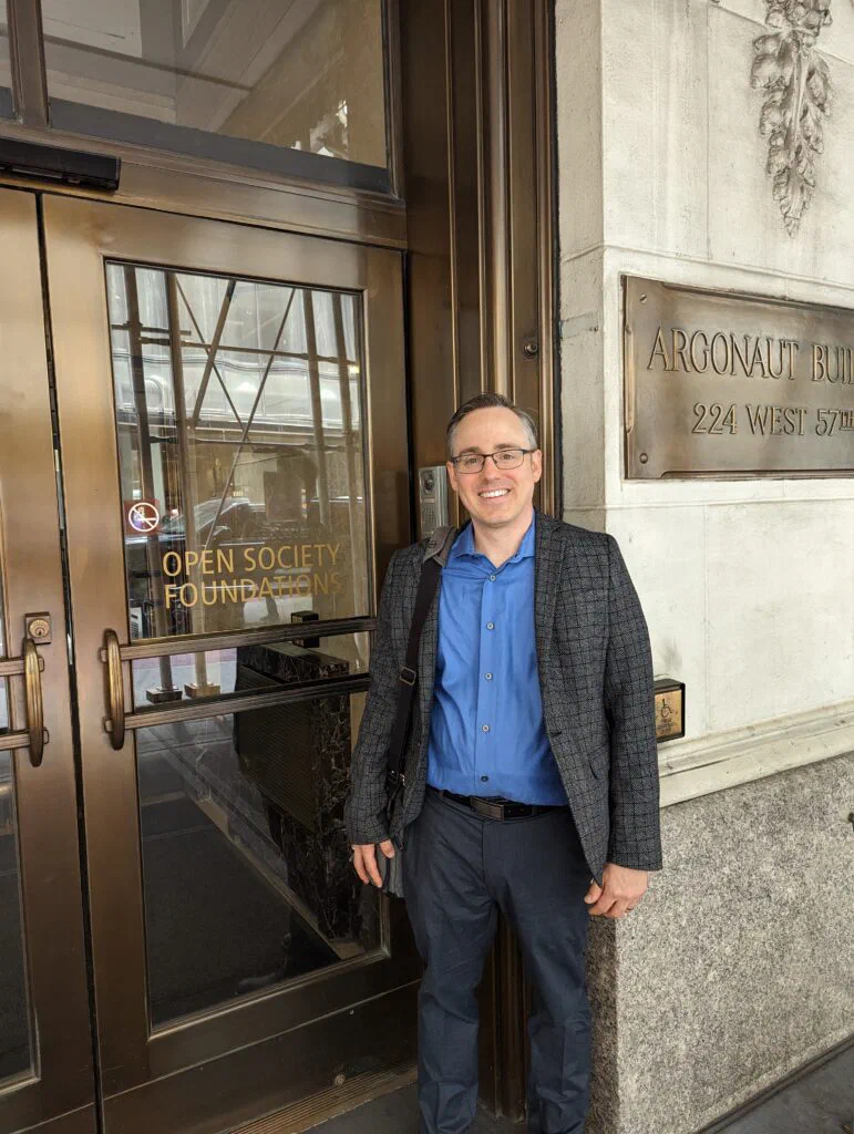 A man, Jared Knowles, stands in front of the Argonaut Building, identified by signage as housing the Open Society Foundations. He is smiling and wearing a blue shirt, grey pants, and a tweed jacket.