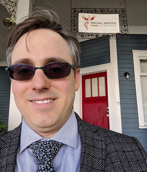 Jared Knowles, a man wearing sunglasses and a suit stands, in front of a blue house with a red door. Hanging above the doorway is a banner for the Racial Justice Action Center, indicating a visit to that organization.