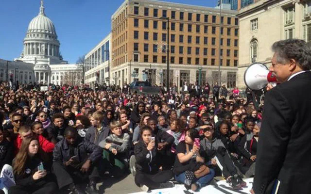 A crowd of students, many wearing purple, fills a plaza in front of a large brick building and the Wisconsin State Capitol. A man with a megaphone addresses the crowd, likely referencing the 2015 protest following Tony Robinson’s death.