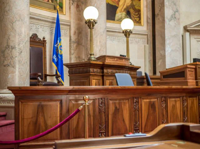 This photograph depicts a portion of a legislative chamber, featuring dark wood paneling, ornate lamps, and a red velvet rope barrier. This is the legislative chambers within the Wisconsin State Capitol.