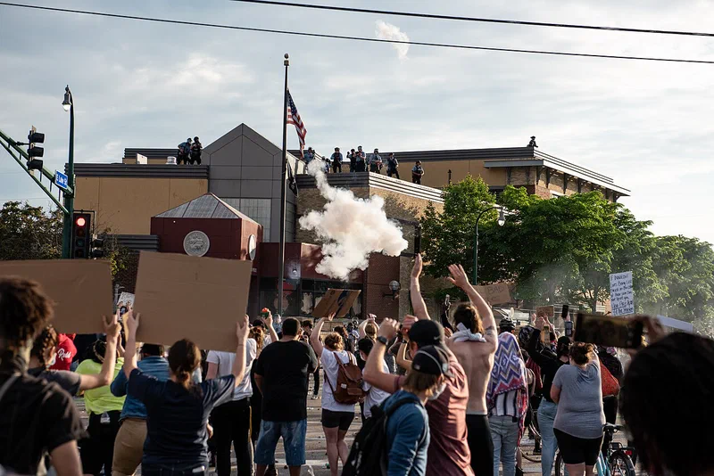 Minneapolis police on the roof of the Third Precinct station fire tear gas toward a crowd of protesters below, many with raised hands and cardboard signs, on May 27, 2020.
