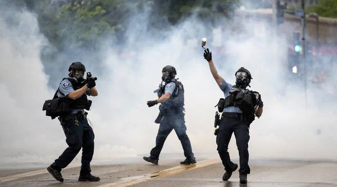 A photograph shows police officers in riot gear deploying tear gas during a protest, with one officer raising his hand. The image illustrates the equipment often associated with police departments, which represents a small portion of their overall budget.