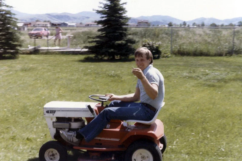 A young man sits on a small, orange riding lawnmower in a grassy yard. The man is smiling and appears to be driving the mower, evoking a sense of childhood fun and adventure.