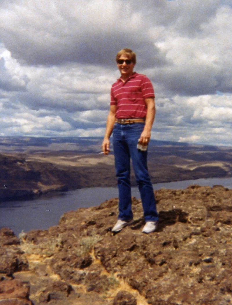 A color photograph shows a man in his 40s, wearing jeans and a striped shirt, standing on a rocky outcrop overlooking a valley. The man is smiling slightly and holding what appears to be a cigarette, embodying his love of exploring new landscapes.