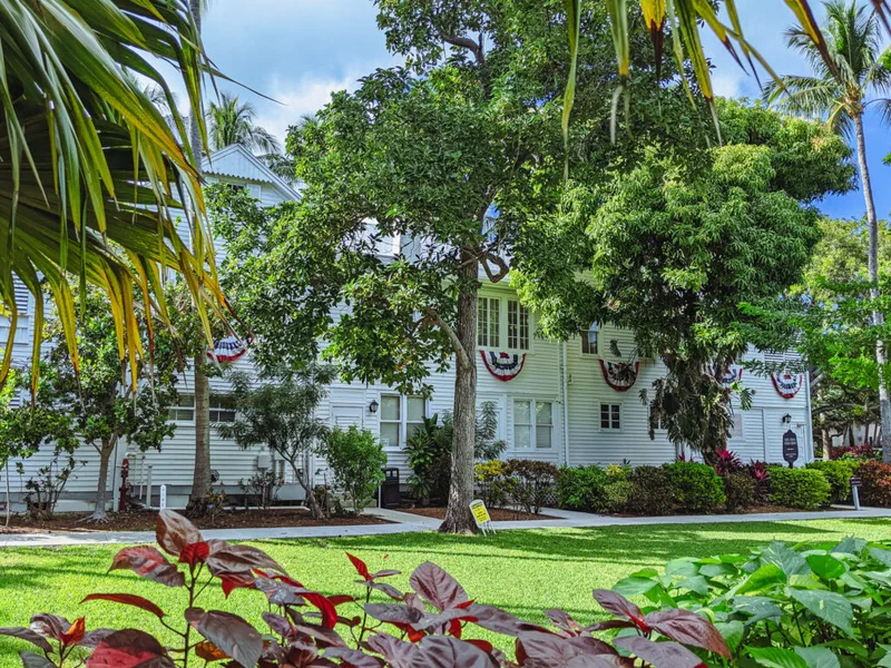 This photograph shows The Little White House in Key West, Florida, decorated with American flags. The white building is framed by lush tropical foliage and a well-manicured lawn, reflecting the setting where President Truman sought decisive counsel.