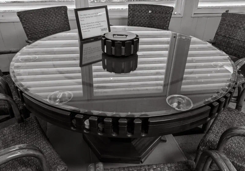 This photograph shows a large, round wooden table with chairs surrounding it, set up for a poker game. A sign on the table reads 'Family during their time in Key West.