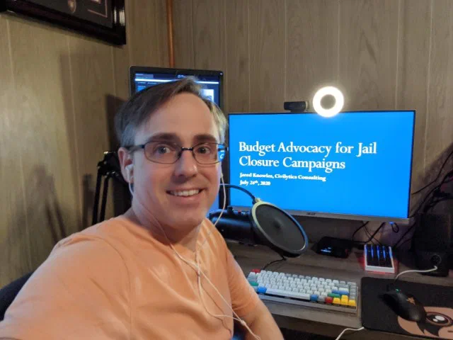 A man with glasses and a headset sits in front of a computer displaying the title 'Budget Advocacy for Jail Closure Campaigns.' The photo, taken in 2020, shows him preparing to go live on Zoom from his home office.